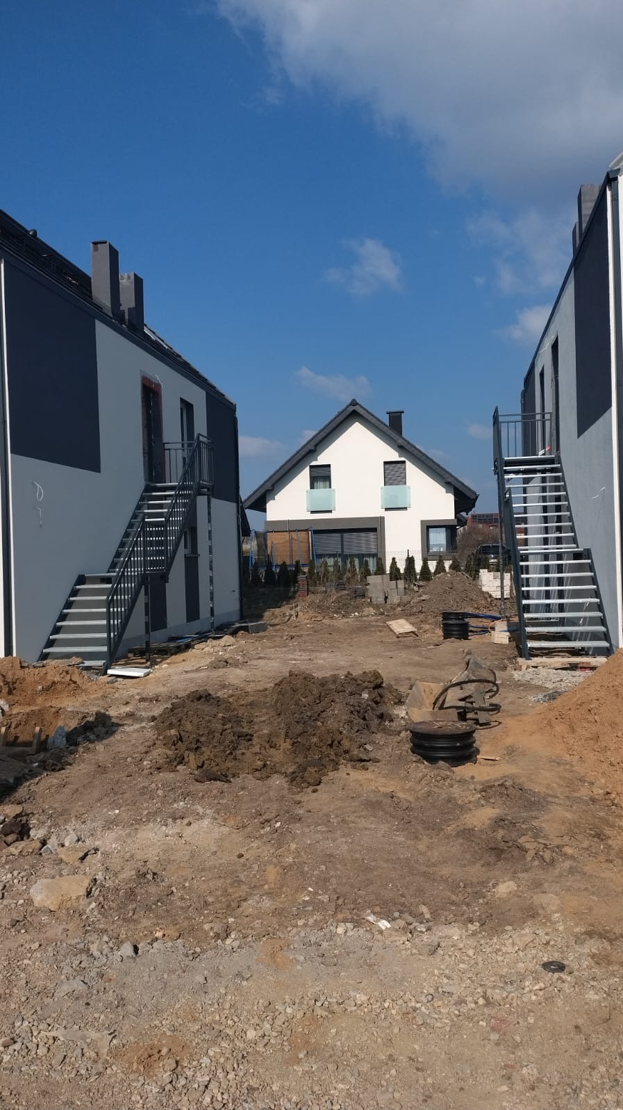 Zewnętrzne schody metalowe z balustradą prowadzące do drzwi na piętrze budynku, widoczne na placu budowy z ziemią i gruzem.