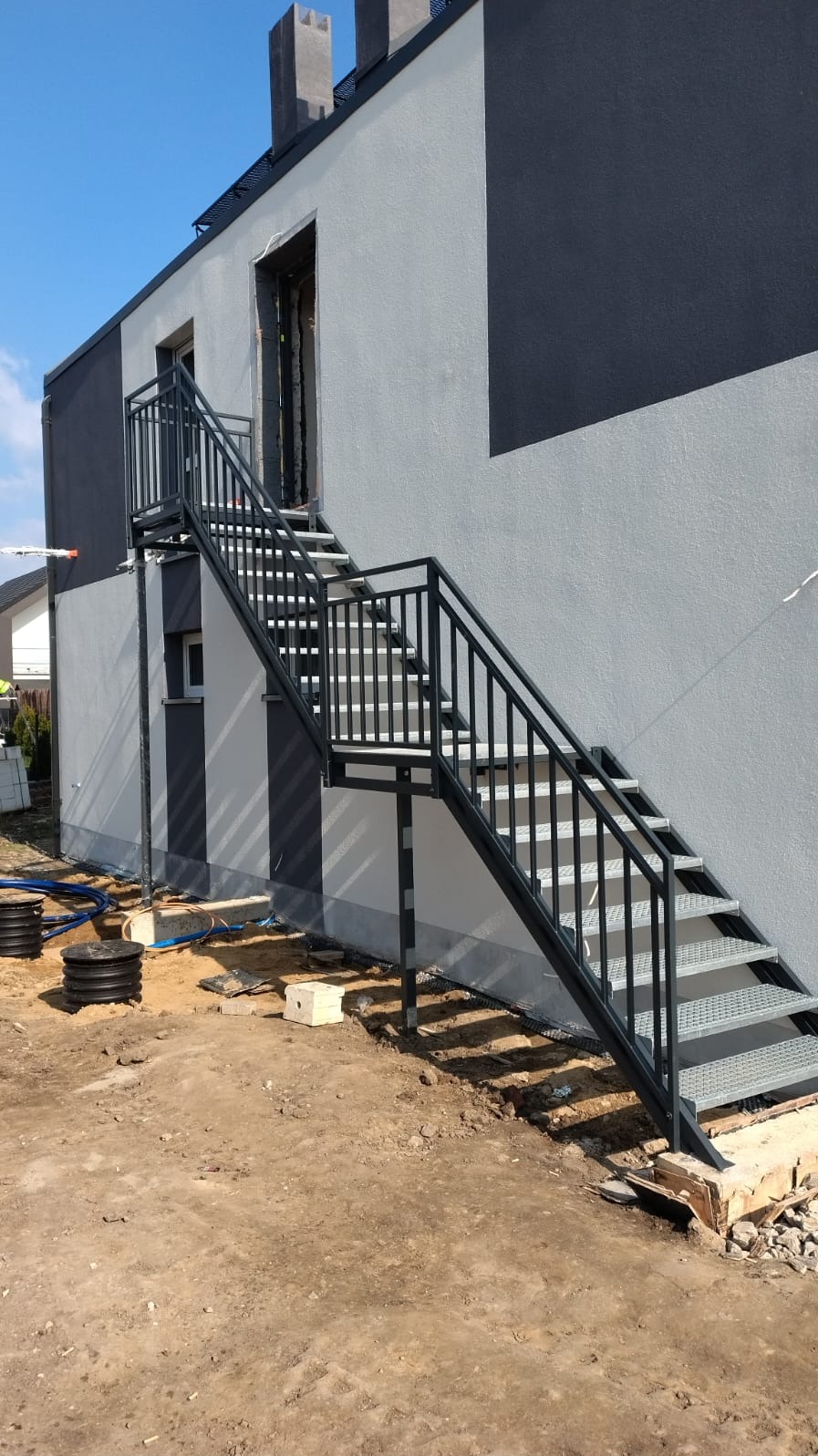 Zewnętrzne metalowe schody z czarną balustradą prowadzące do drzwi na piętrze budynku o szaro-czarnej elewacji, widoczne prace ziemne wokół.