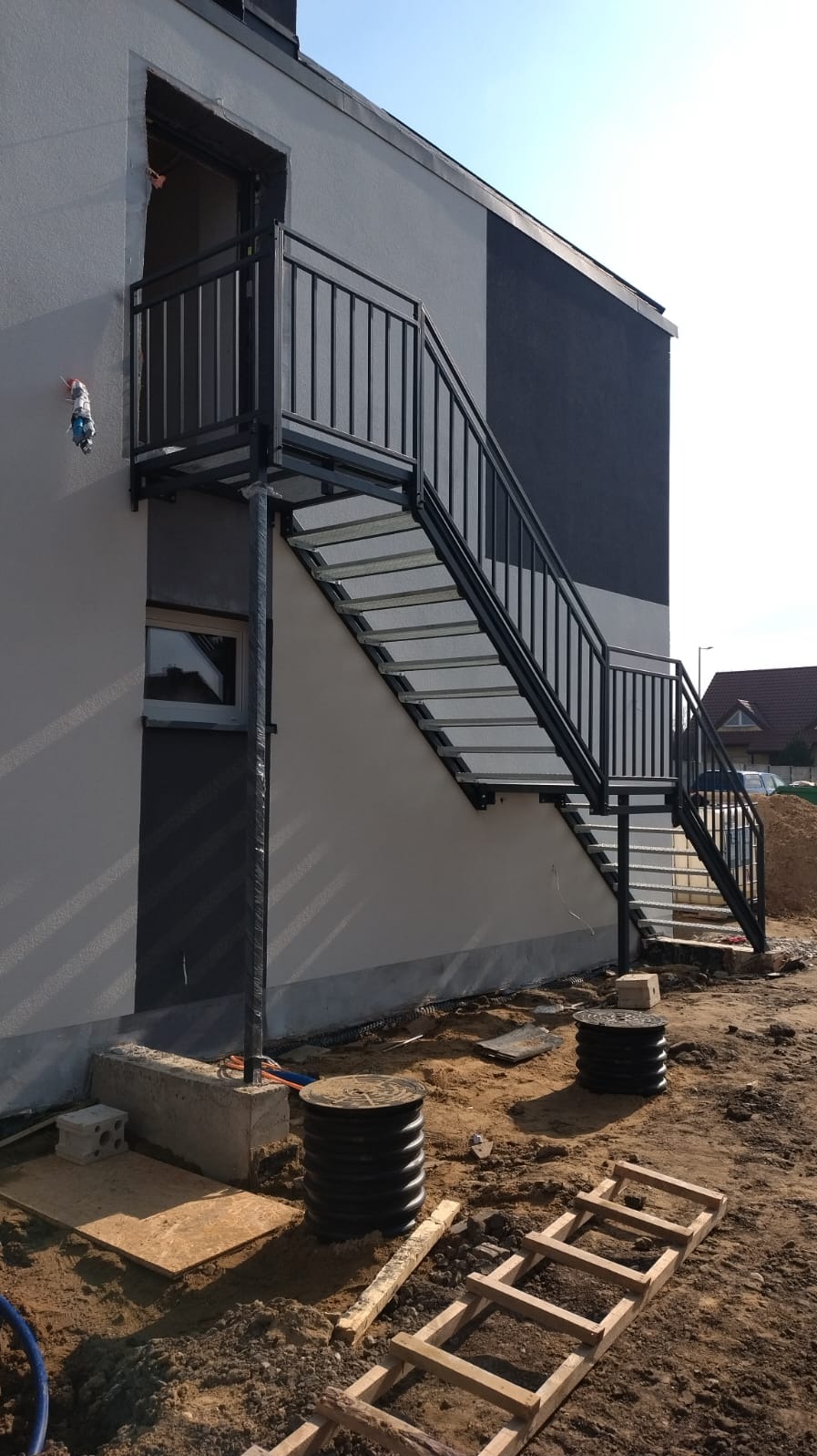 Zewnętrzne schody metalowe z balustradą prowadzące do drzwi na piętrze, widoczne elementy konstrukcyjne i teren budowy.