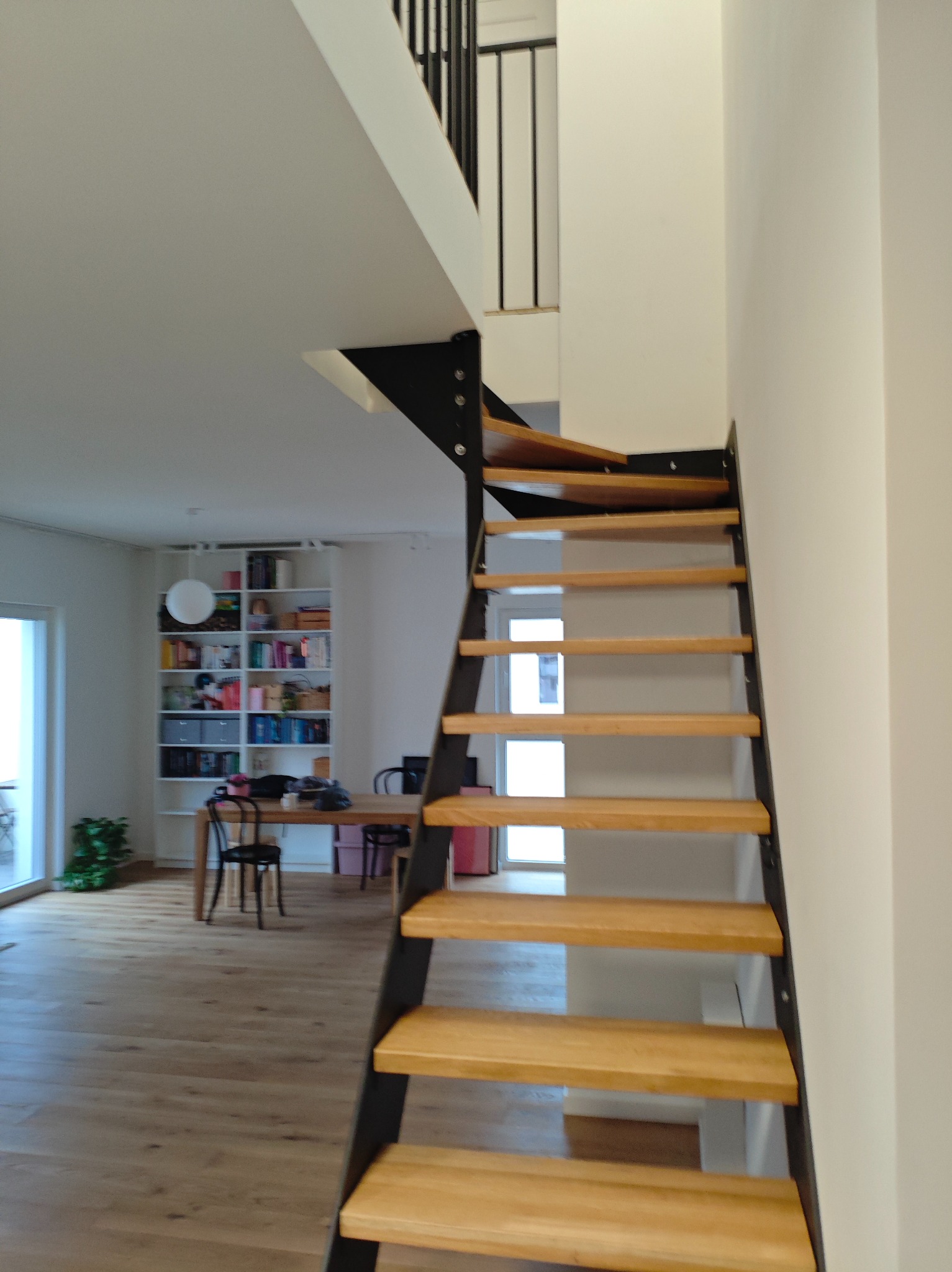Nowoczesne schody z drewnianymi stopniami i czarną metalową konstrukcją, widok z dołu, w tle jasne wnętrze z regałem na książki.