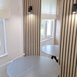 Usługi Remontowo Budowlane Paweł Gacek - Nowoczesne wnętrze z okładziną ścienną z drewnianych paneli, okrągłym stołem z szarą marmurkową powierzchnią i czarnymi nogami, oraz minimalistycznym oświetleniem punktowym.