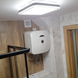 Usługi Remontowo Budowlane Paweł Gacek - Nowoczesna łazienka z marmurowymi płytkami, podwieszanym bojlerem Ariston, minimalistycznym oświetleniem sufitowym i fragmentem czarnej kabiny prysznicowej.