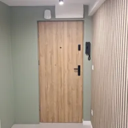 Nowoczesne wejście do mieszkania z drewnianymi panelami ściennymi, drzwi wejściowe z okleiną drewnopodobną i czarnymi okuciami, ściana pomalowana na stonowany odcień zieleni.