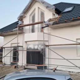 Dom jednorodzinny w trakcie prac elewacyjnych, zabezpieczony folią i rusztowaniem, widoczny fragment srebrnego samochodu na pierwszym planie.