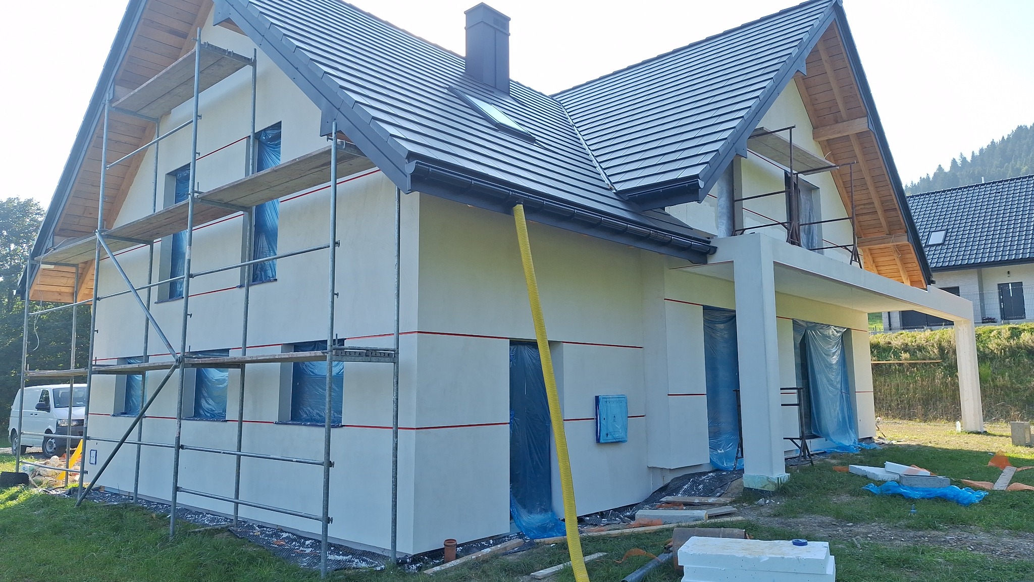 Nowo otynkowany dom jednorodzinny w trakcie wykańczania, z rusztowaniami, zabezpieczonymi oknami i drzwiami, widoczne elementy ocieplenia i wentylacji.