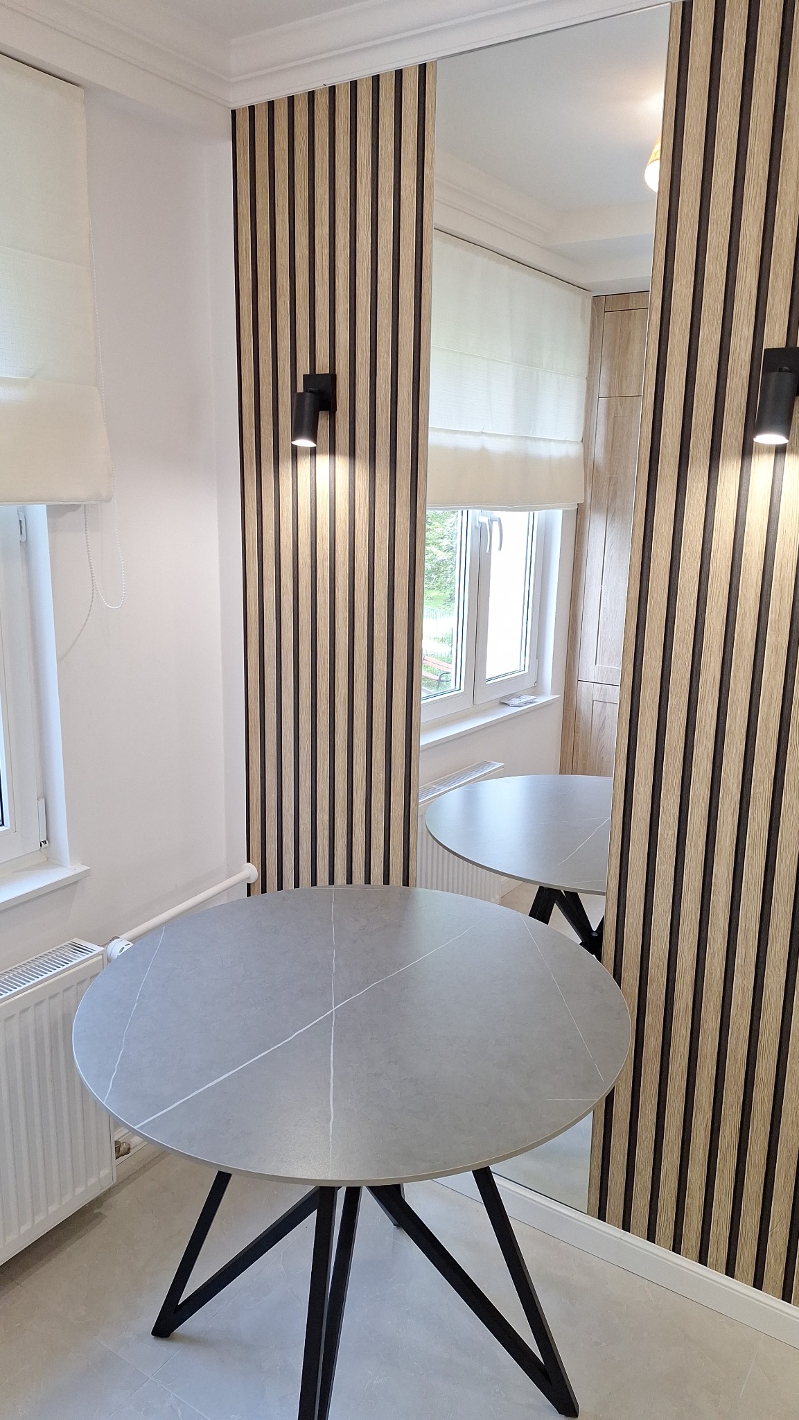 Nowoczesne wnętrze z okładziną ścienną z drewnianych paneli, okrągłym stołem z szarą marmurkową powierzchnią i czarnymi nogami, oraz minimalistycznym oświetleniem punktowym.