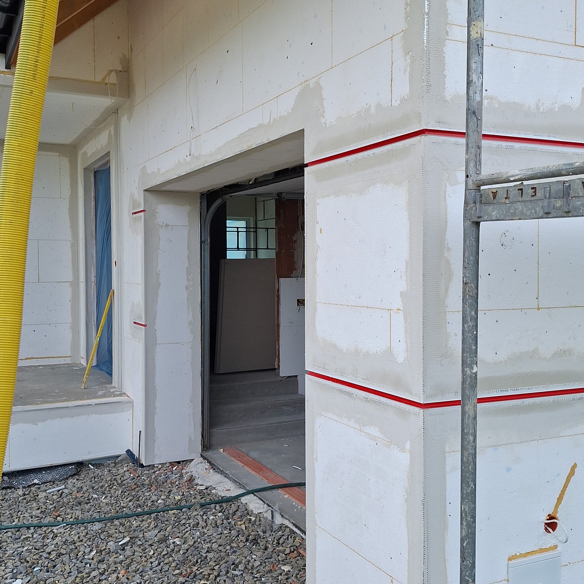 Budynek w Olszówce w trakcie ocieplania, widoczne białe płyty izolacyjne z zaznaczonymi czerwoną taśmą liniami, żółta rura wentylacyjna i fragment rusztowania.