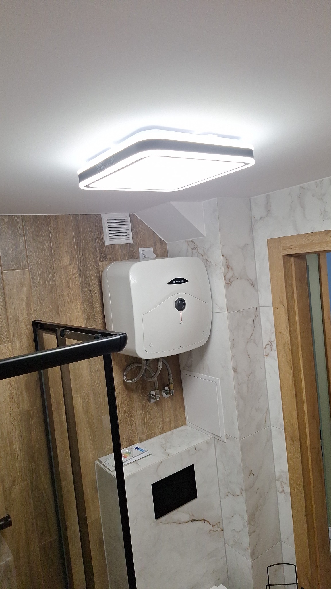 Nowoczesna łazienka z marmurowymi płytkami, podwieszanym bojlerem Ariston, minimalistycznym oświetleniem sufitowym i fragmentem czarnej kabiny prysznicowej.