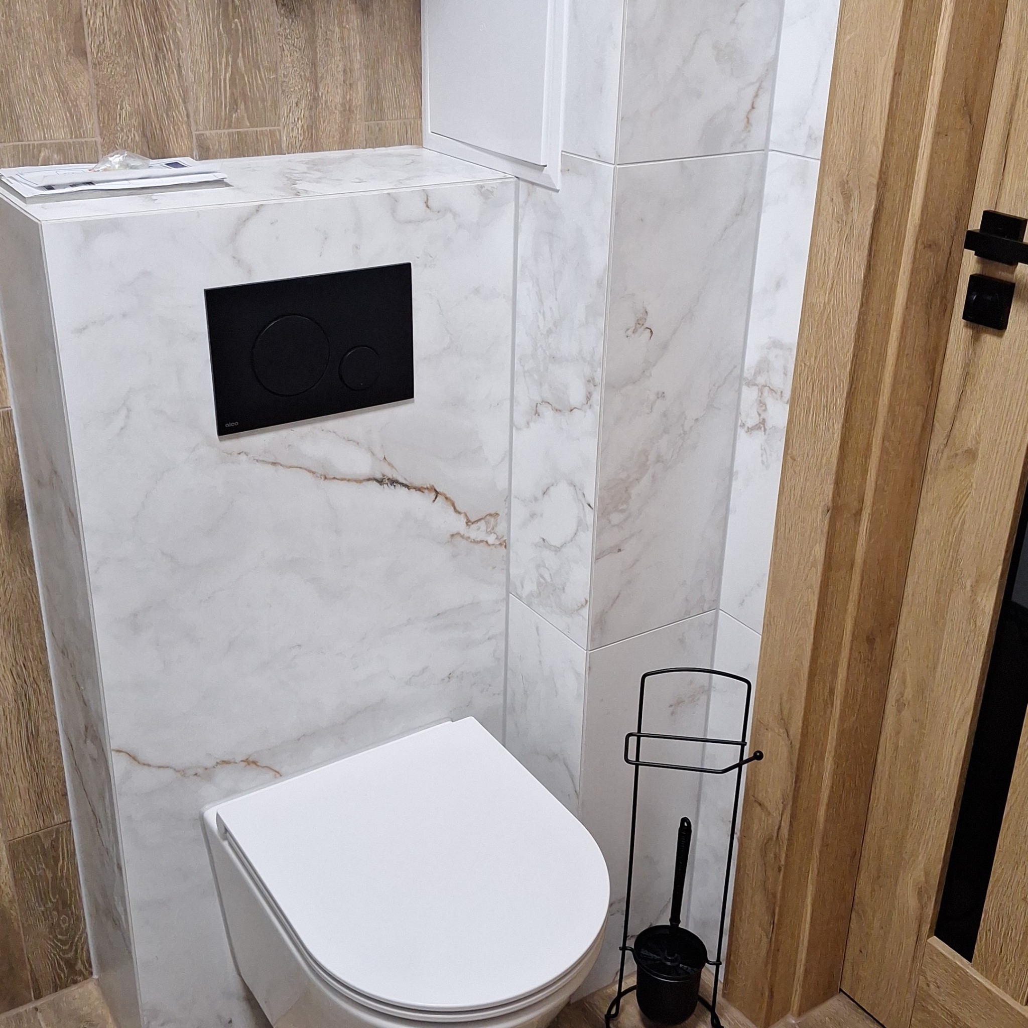 Nowoczesna toaleta podwieszana z czarnym przyciskiem spłukującym na tle płytek imitujących marmur, obok drewniane drzwi i czarny stojak na szczotkę toaletową.
