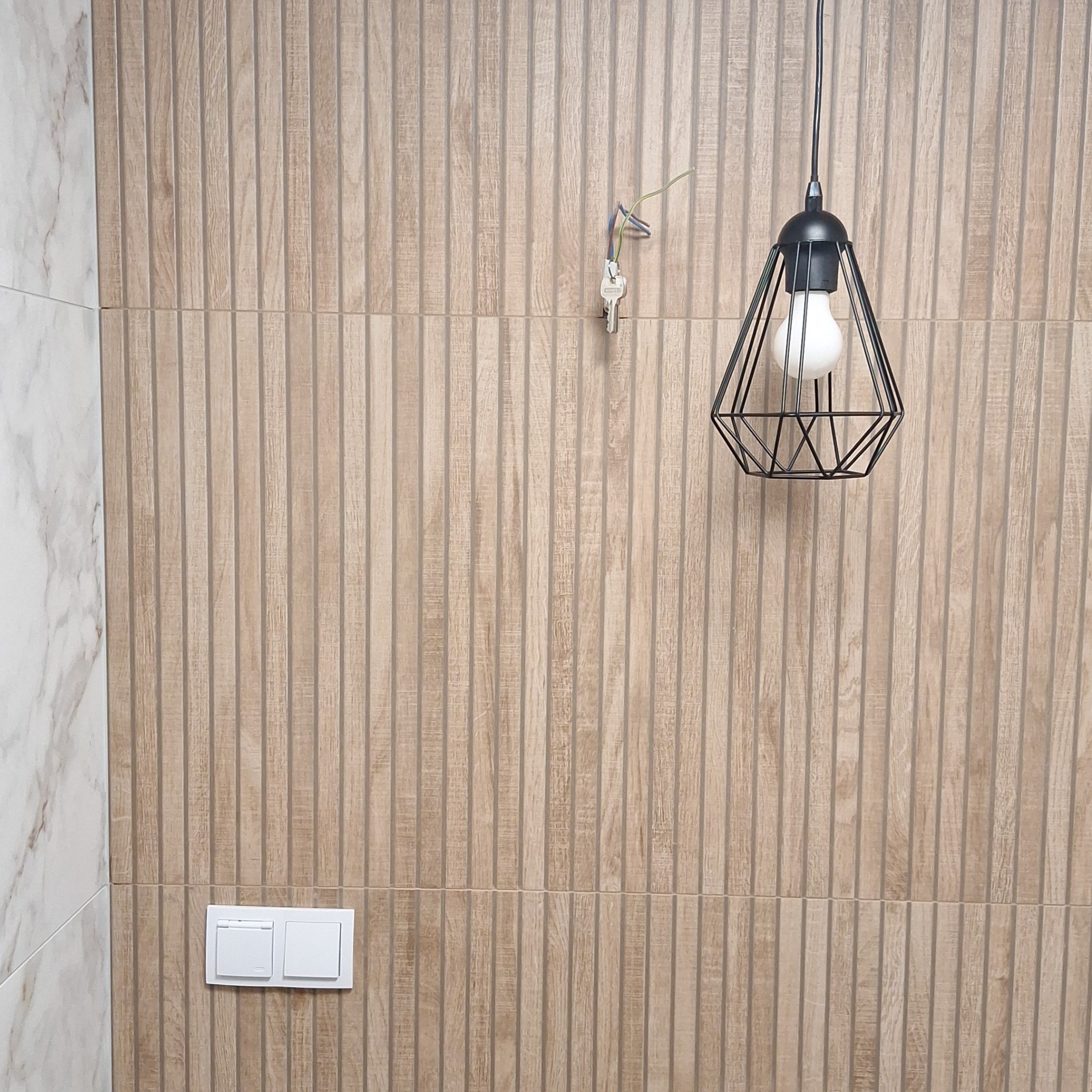 Ściana wyłożona panelami imitującymi drewno z włącznikiem światła i wiszącą lampą drucianą z żarówką Edisona, obok fragment ściany wyłożonej marmurem.