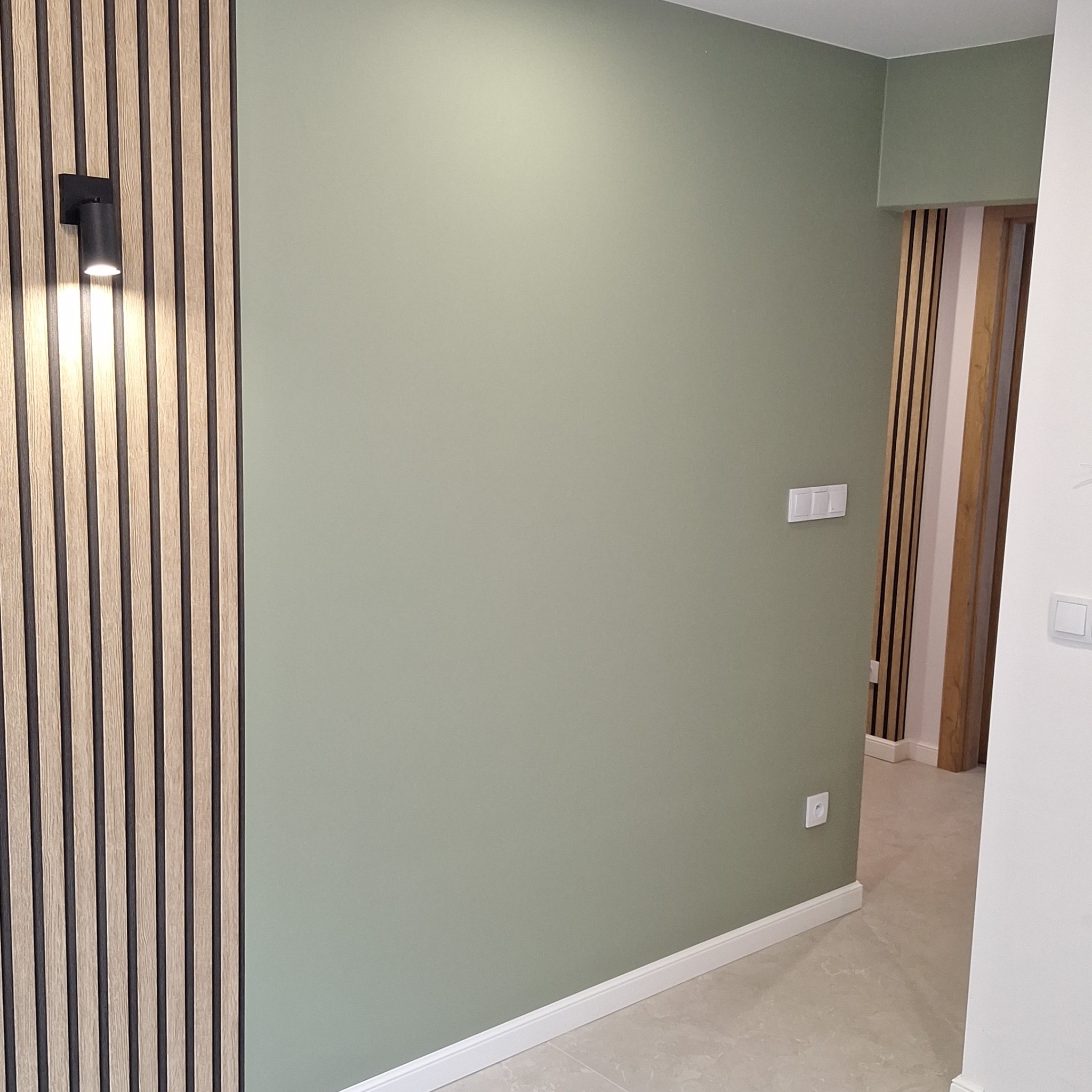 Ściana pomalowana na oliwkowy kolor z dekoracyjnymi panelami ściennymi z pionowymi listwami w kolorze drewna i czerni oraz oświetleniem punktowym, widoczna podłoga z płytek w odcieniach szarości...