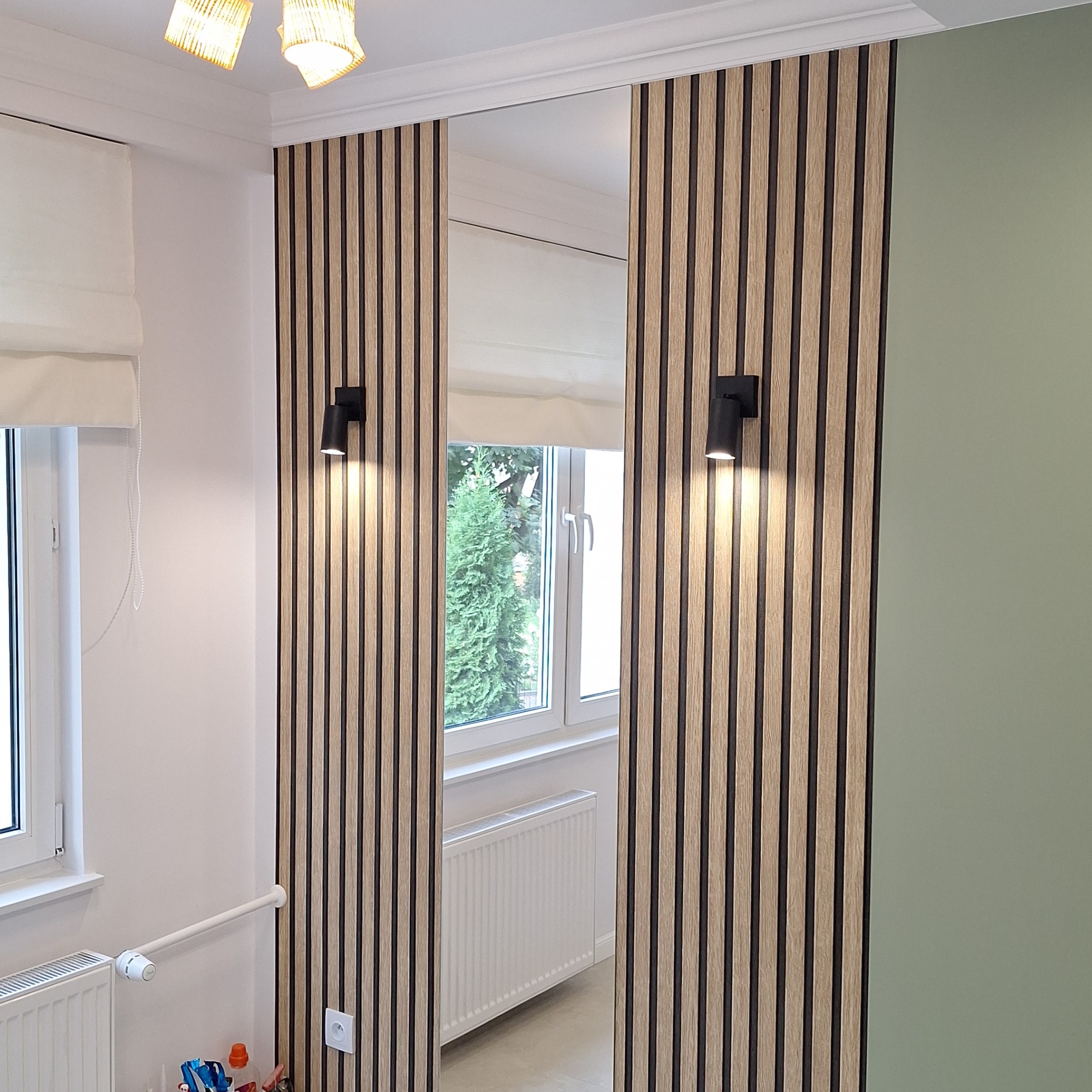 Panel ścienny z drewnianych listew z czarnymi wstawkami, oświetlony czarnymi kinkietami, z widocznym fragmentem okna i grzejnika w odbiciu lustra.