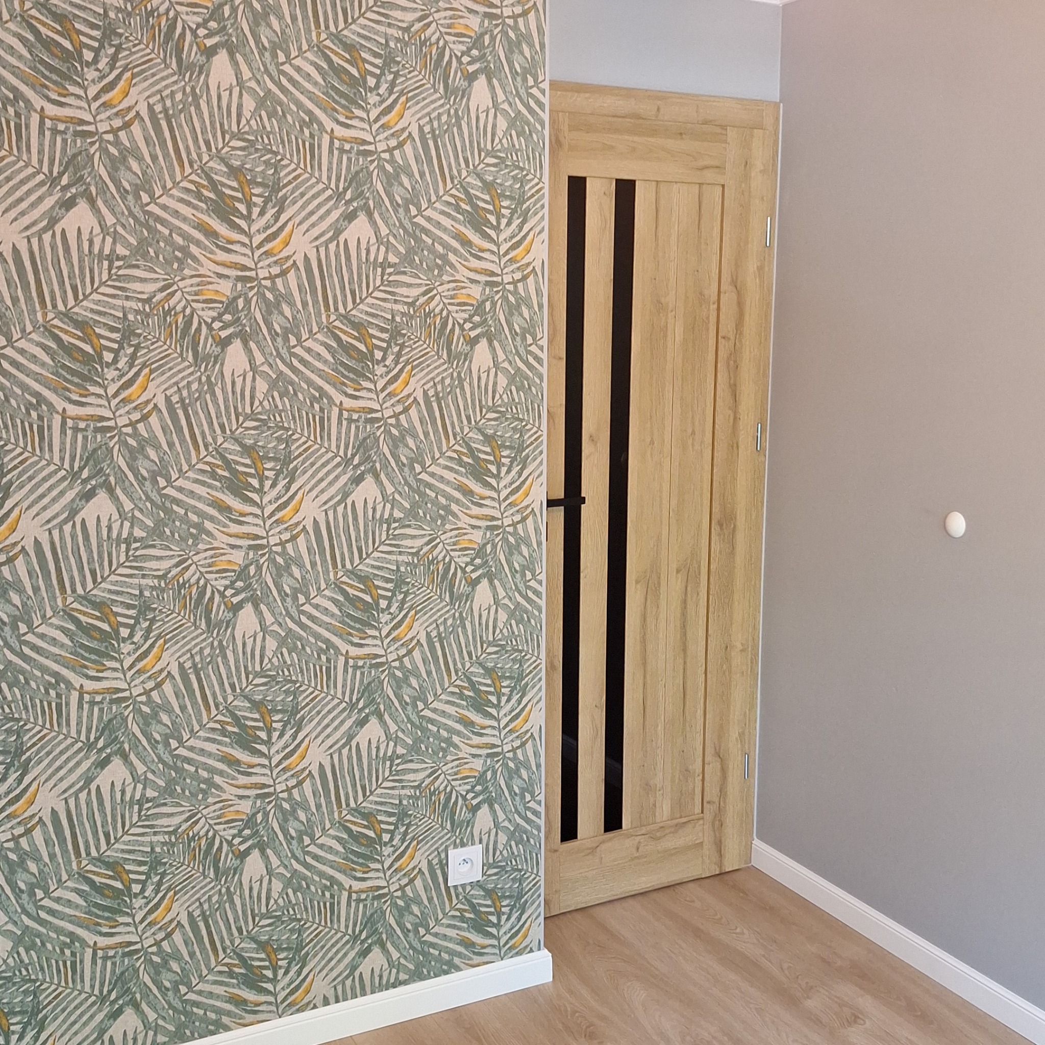 Ściana z tapetą w tropikalne liście obok drewnianych drzwi z czarnymi wstawkami, widoczny fragment podłogi i ściany pomalowanej na szaro.