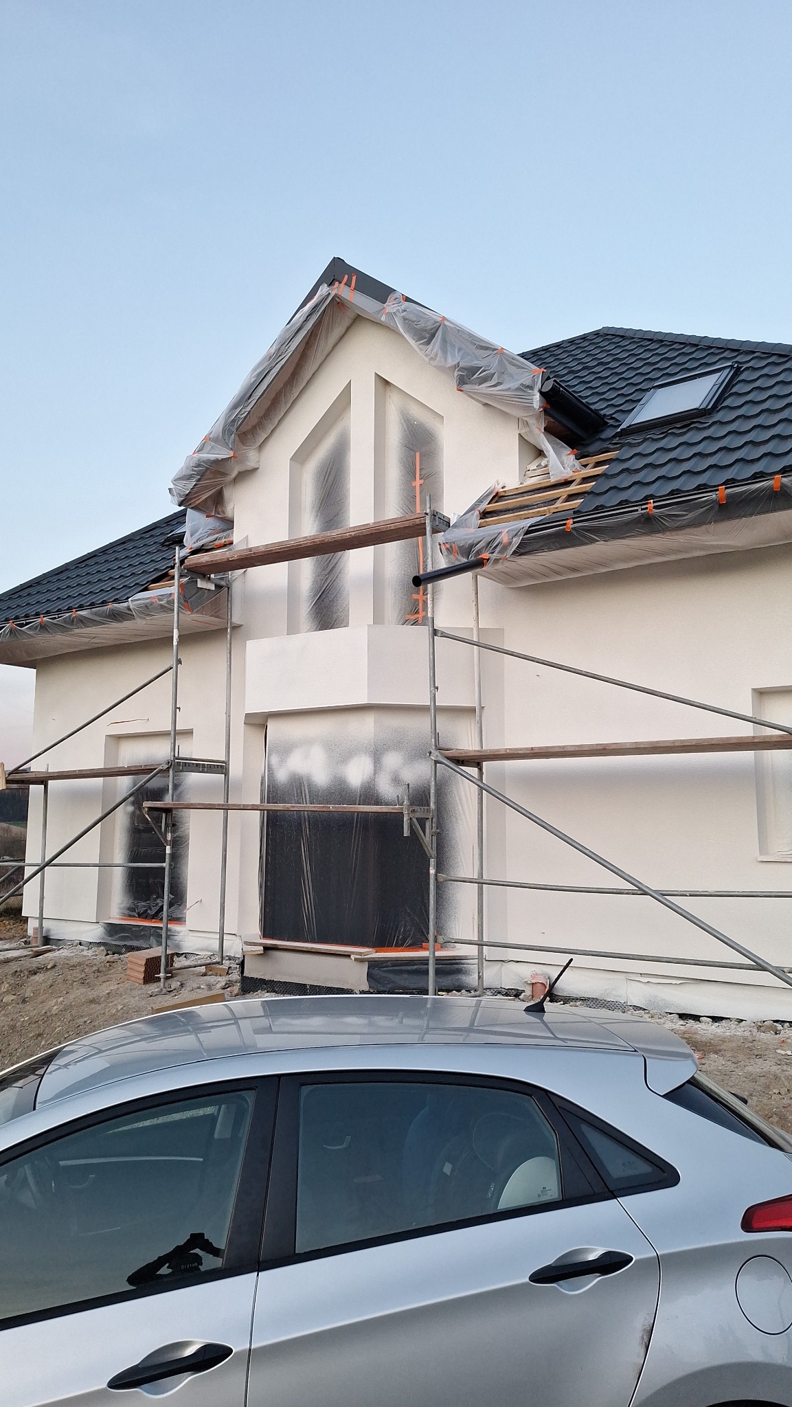 Dom jednorodzinny w trakcie prac elewacyjnych, zabezpieczony folią i rusztowaniem, widoczny fragment srebrnego samochodu na pierwszym planie.