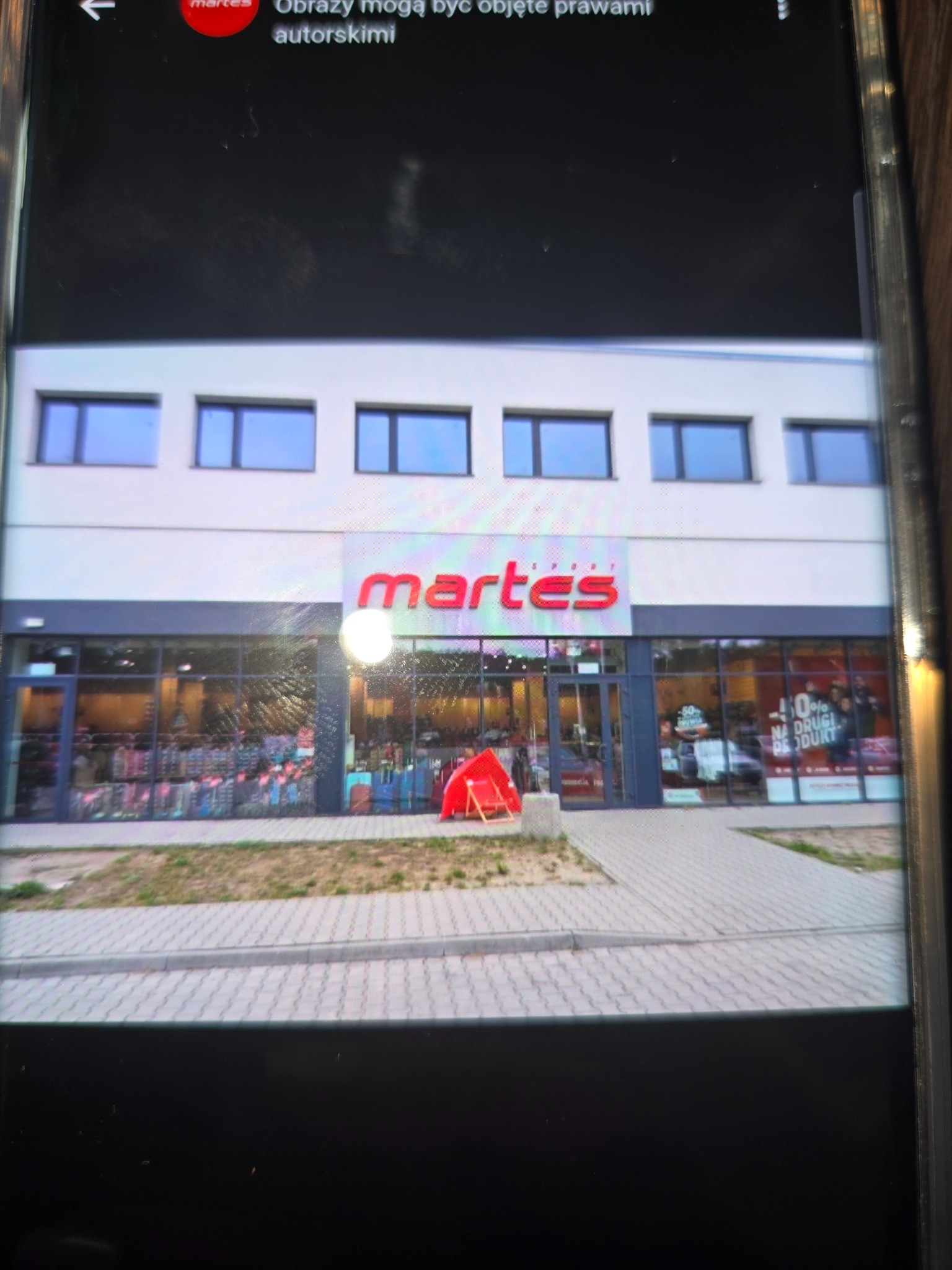 Fasada sklepu Martes Sport z dużymi oknami wystawowymi i chodnikiem z kostki brukowej. Widoczny neon z nazwą sklepu i promocje na szybach.