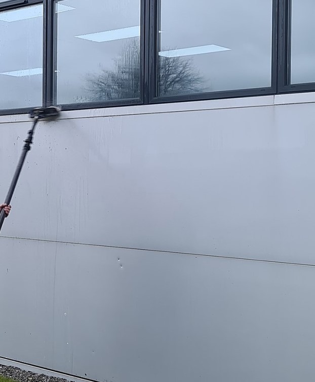 Mycie elewacji budynku z użyciem szczotki na teleskopowym wysięgniku. Widoczne ślady wody na elewacji i odbicie drzewa w oknach.