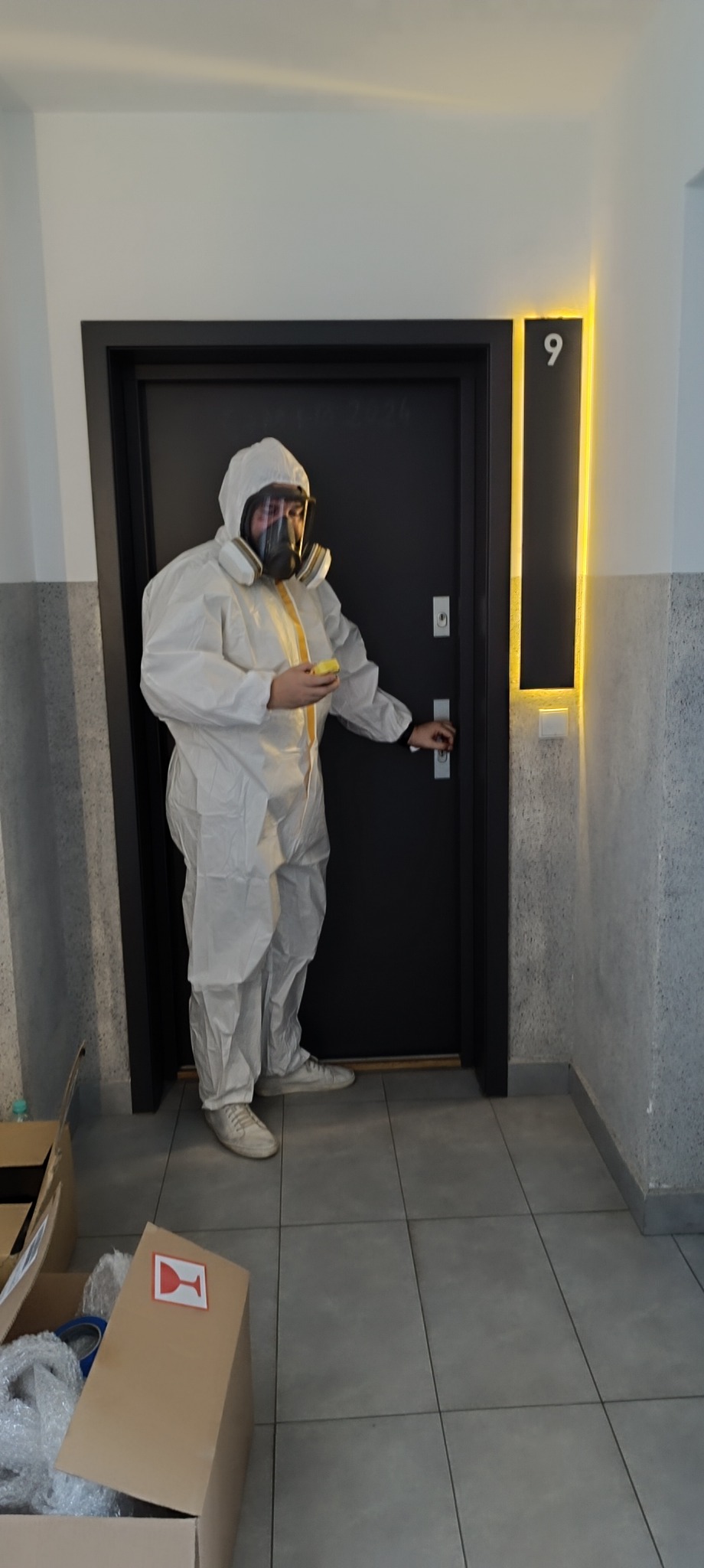 Pracownik w kombinezonie ochronnym z maską gazową, trzymający gąbkę, stoi przed drzwiami z numerem 9, w tle widoczne kartony.