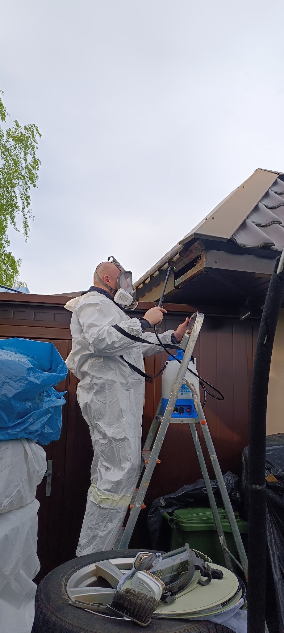 Pracownik w białym kombinezonie ochronnym i masce, stojący na aluminiowej drabinie, opryskuje preparatem drewnianą podbitkę dachu przy pomocy lancy z niebieskiego opryskiwacza ciśnieniowego.