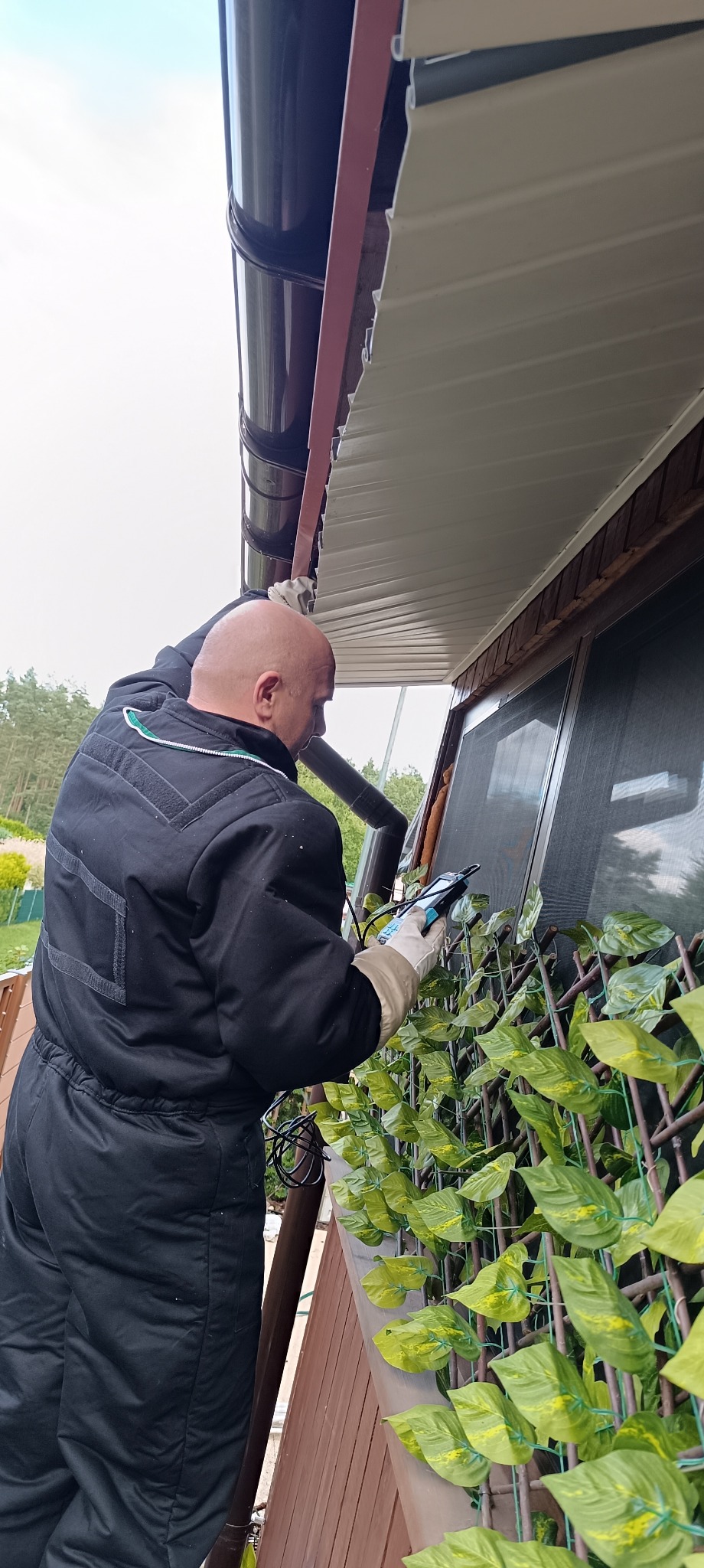 Mężczyzna w kombinezonie sprawdza stan okna za pomocą elektronicznego urządzenia pomiarowego, widoczne rynny i sztuczne pnącze na balkonie.