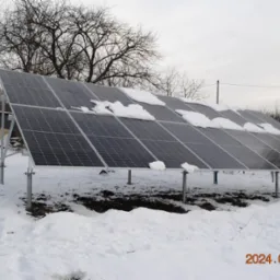 Panele fotowoltaiczne pokryte śniegiem w zimowej scenerii Kryspinowa, widoczne metalowe podpory i częściowo odśnieżona ziemia.