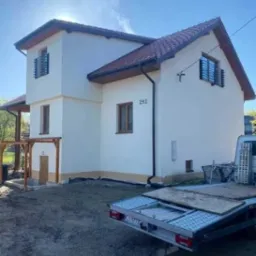 Dwupiętrowy dom z beżową elewacją i czerwoną dachówką, widoczny z boku, z numerem 292 na ścianie, częściowo zasłonięty przez przyczepę transportową na niezagospodarowanej działce.