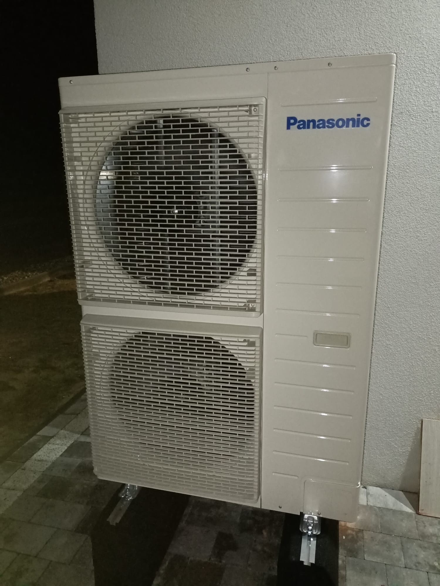 Zewnętrzna jednostka klimatyzacji Panasonic z dwoma wentylatorami, zamontowana przy szarej ścianie budynku, widoczna na tle ciemności.