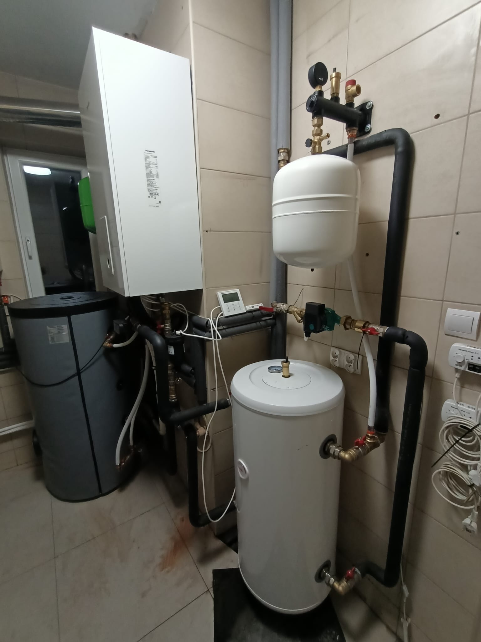 Kompaktowa kotłownia w pomieszczeniu z beżowymi płytkami, zawierająca biały piec, szary zbiornik akumulacyjny, biały zasobnik CWU, czarne rury instalacyjne oraz osprzęt pomiarowy i sterujący.
