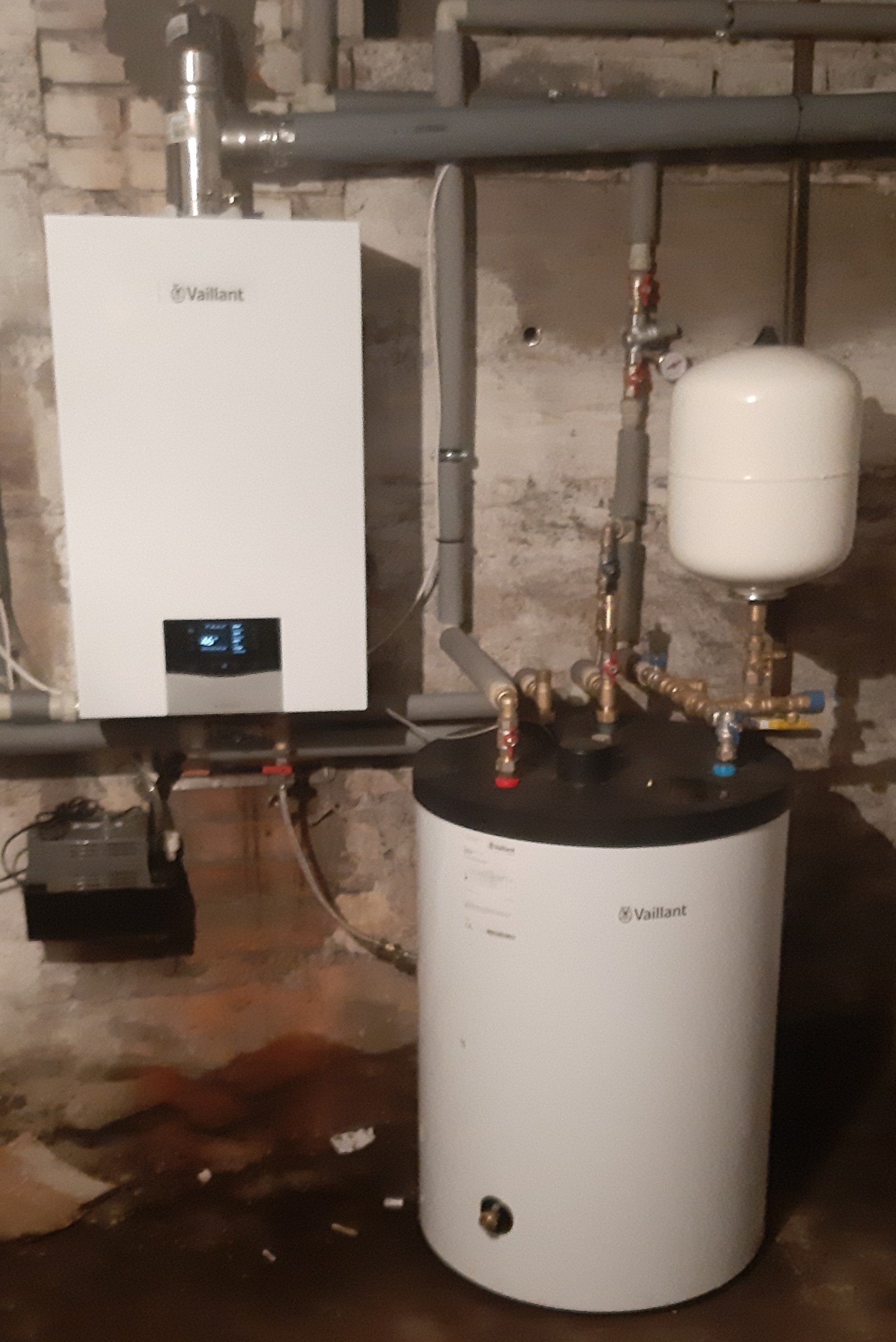 Biały piec gazowy Vaillant wiszący na ścianie z podłączonymi rurami, obok biały zasobnik ciepłej wody z czarnym blatem i zbiornikiem wyrównawczym w piwnicy.