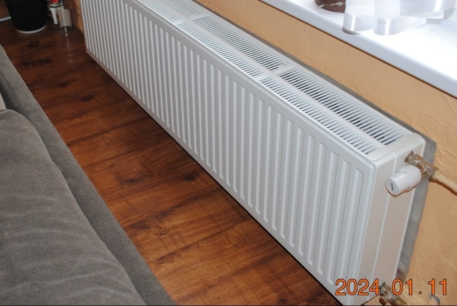 Biały, żebrowany grzejnik panelowy zamontowany przy ścianie, widoczny zawór termostatyczny i fragment rury instalacji grzewczej, na podłodze panele imitujące drewno.