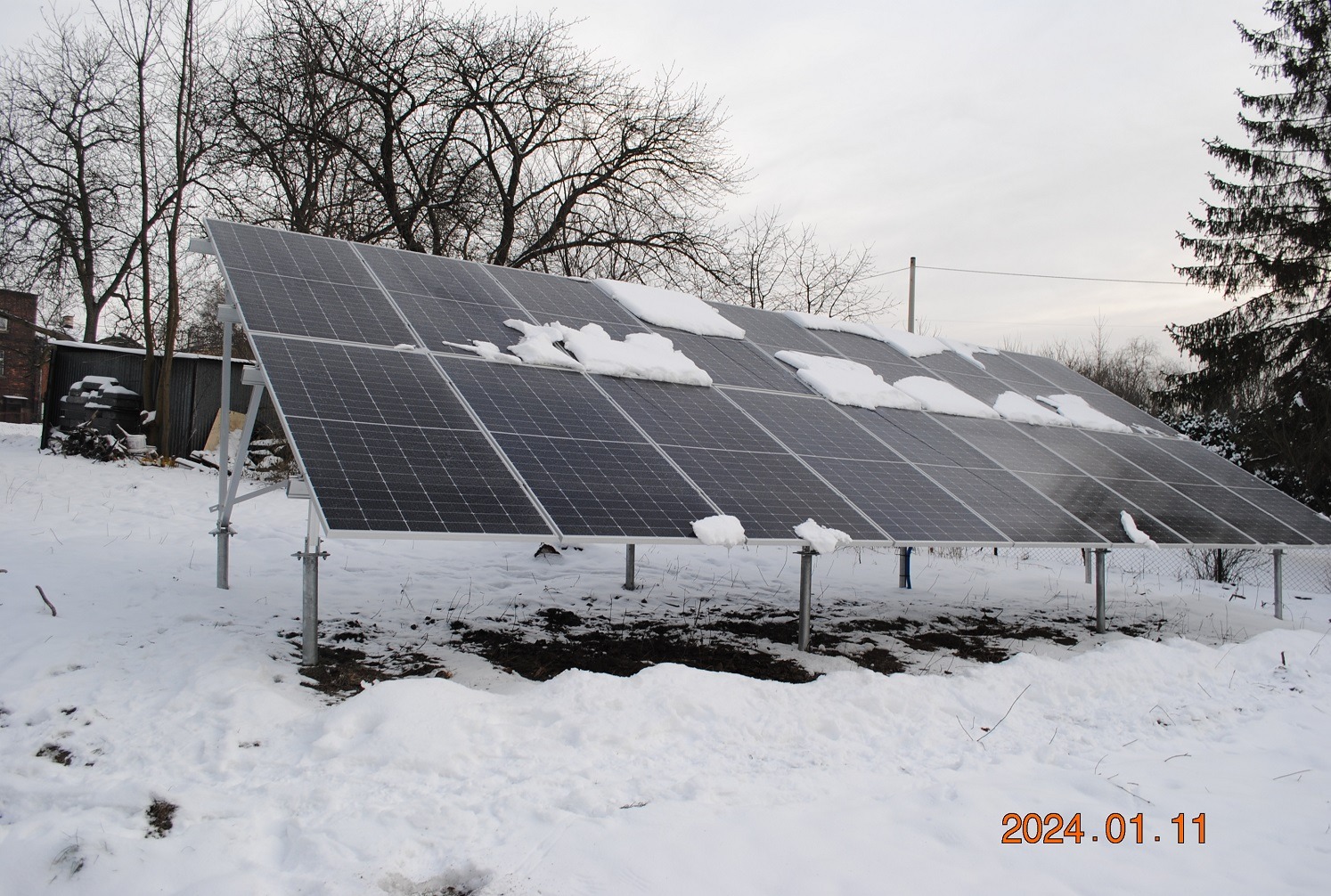 Panele fotowoltaiczne pokryte śniegiem w zimowej scenerii Kryspinowa, widoczne metalowe podpory i częściowo odśnieżona ziemia.