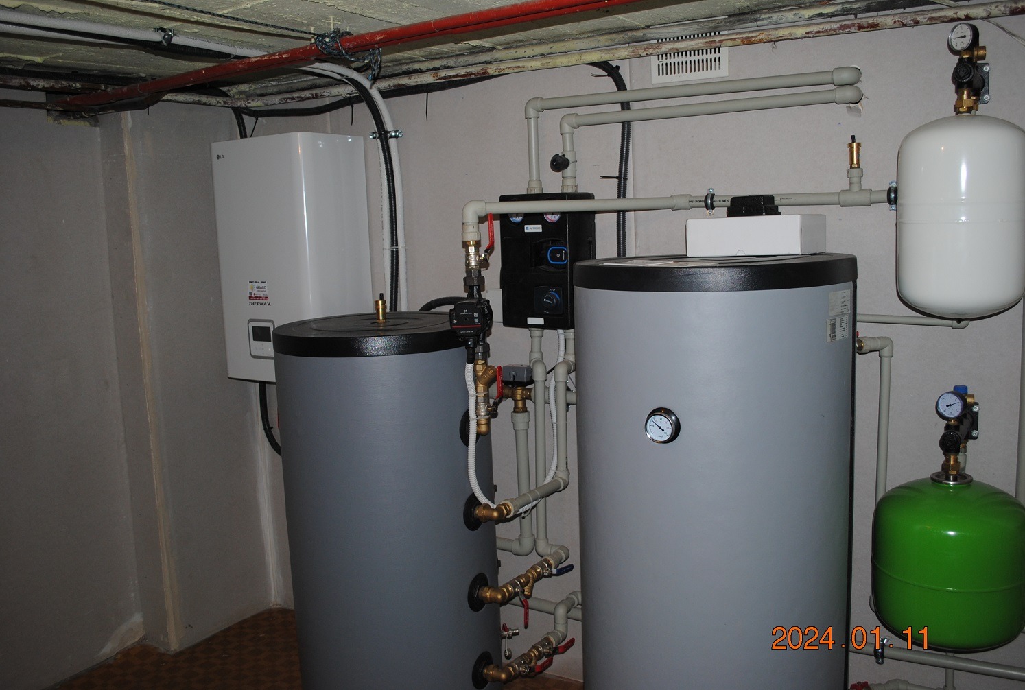 Dwa szare zbiorniki akumulacyjne ciepła z podłączoną instalacją hydrauliczną, obok biały piec wiszący LG Therma V, całość w piwnicy z rurami pod sufitem.
