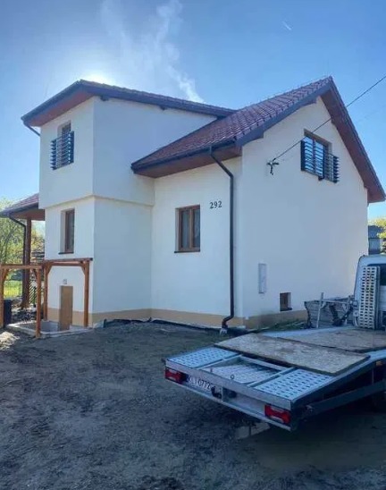 Dwupiętrowy dom z beżową elewacją i czerwoną dachówką, widoczny z boku, z numerem 292 na ścianie, częściowo zasłonięty przez przyczepę transportową na niezagospodarowanej działce.