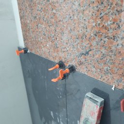 Mateusz Górski - Układanie ciemnoszarych płytek z wykorzystaniem poziomicy i pomarańczowych klipsów poziomujących na ścianie z czerwonego, nakrapianego granitu.