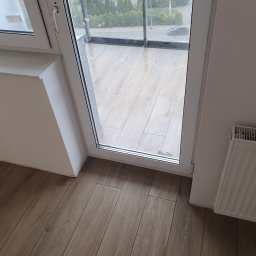 Mateusz Górski - Balkon z drewnopodobnymi płytkami podłogowymi, widok z wnętrza mieszkania przez otwarte, białe drzwi balkonowe na zewnątrz.