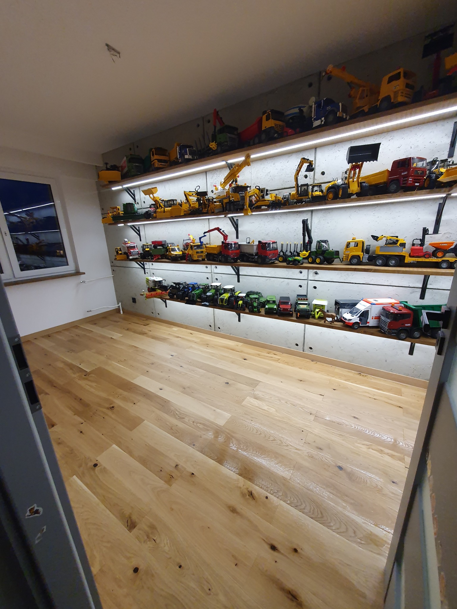 Półki z podświetleniem LED prezentują kolekcję modeli maszyn budowlanych i rolniczych w pokoju z jasną, drewnianą podłogą.