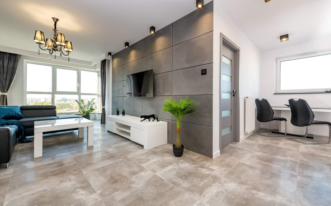 Salon z szarymi płytkami podłogowymi imitującymi kamień, ściana z betonowych paneli, czarna kanapa z turkusową poduszką, biała szafka RTV z dekoracjami, oświetlenie punktowe i żyrandol.