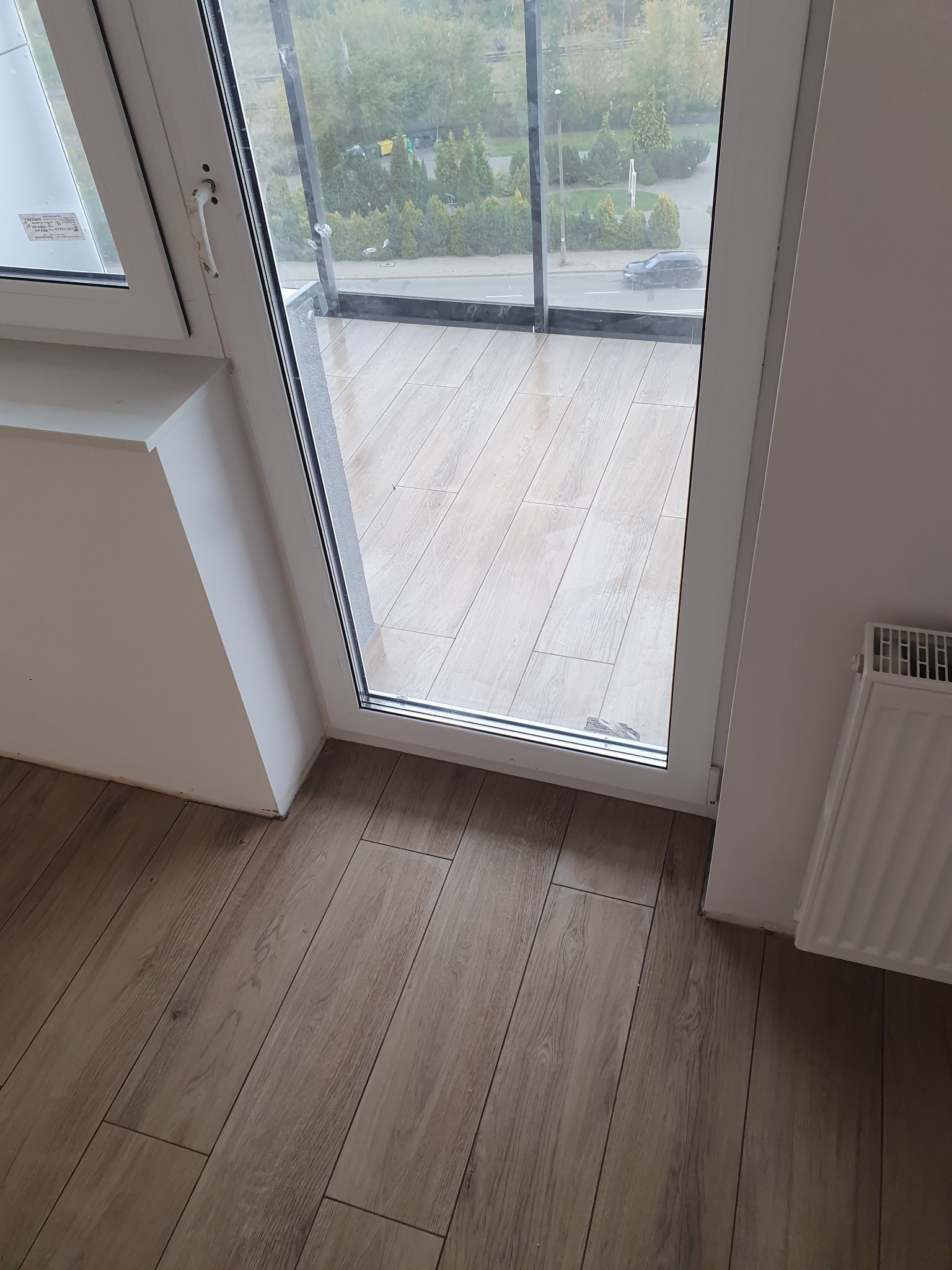 Balkon z drewnopodobnymi płytkami podłogowymi, widok z wnętrza mieszkania przez otwarte, białe drzwi balkonowe na zewnątrz.