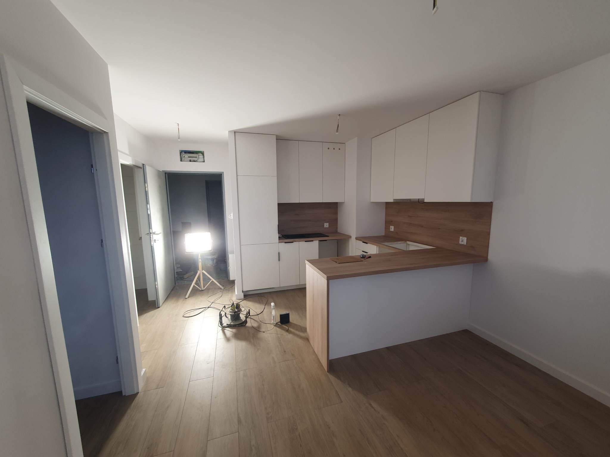 Nowoczesna kuchnia z drewnianym blatem i białymi szafkami, oświetlona lampą budowlaną podczas prac wykończeniowych, widoczne panele podłogowe imitujące drewno.