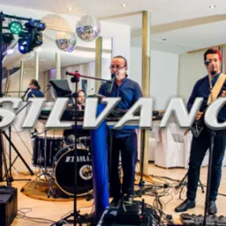 Zespół muzyczny składający się z basisty, klawiszowca/wokalisty, gitarzysty z czerwonymi włosami i saksofonisty występuje na scenie z oświetleniem i kulami dyskotekowymi.