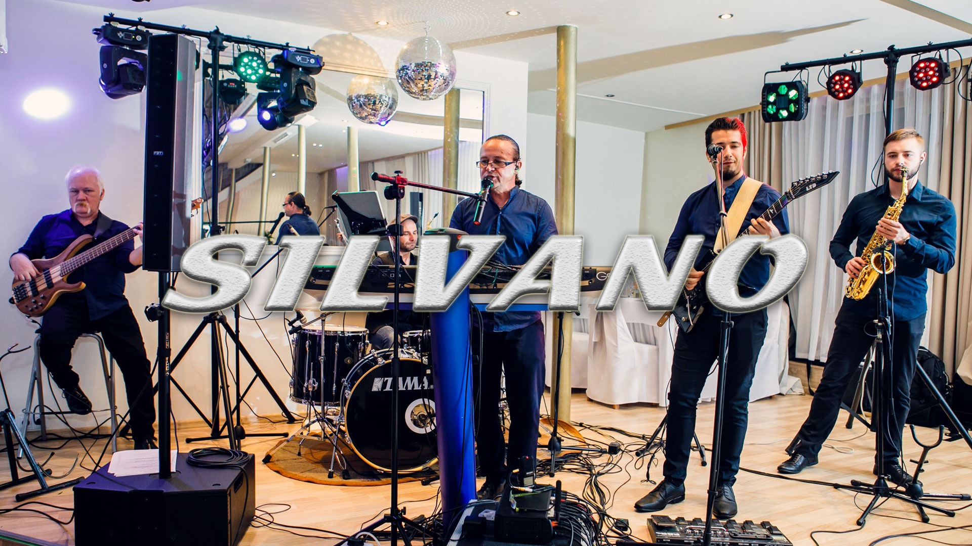 Zespół muzyczny składający się z basisty, klawiszowca/wokalisty, gitarzysty z czerwonymi włosami i saksofonisty występuje na scenie z oświetleniem i kulami dyskotekowymi.