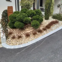 Ogród przydomowy z kulistymi krzewami, kamieniami ozdobnymi i tabliczką powitalną w rustykalnym stylu.