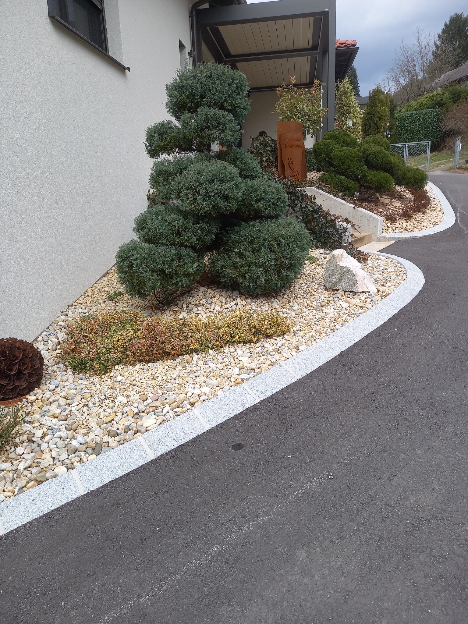 Aranżacja ogrodu przydomowego z ozdobnymi kamieniami, formowanymi krzewami i roślinami okrywowymi, obrzeże z kostki granitowej oddzielające rabatę od asfaltowej drogi dojazdowej.