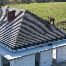 Budexin - Budowa domu jednorodzinnego: montaż łat na dachu, widoczny komin z napisem 'MAXX PLUS', okno dachowe, oraz pracownik w żółtej bluzie na tle zielonego pola.