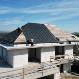 Budowa domu jednorodzinnego: etap montażu czarnego pokrycia dachowego na drewnianej konstrukcji więźby, widoczni dwaj pracownicy na dachu oraz białe ściany z betonu komórkowego parteru, drabina...