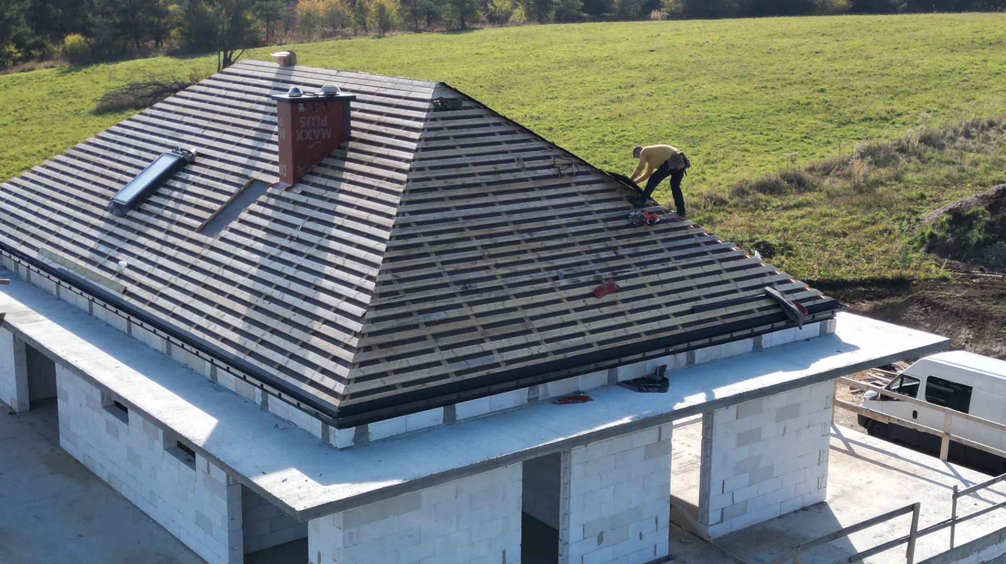Budowa domu jednorodzinnego: montaż łat na dachu, widoczny komin z napisem 'MAXX PLUS', okno dachowe, oraz pracownik w żółtej bluzie na tle zielonego pola.