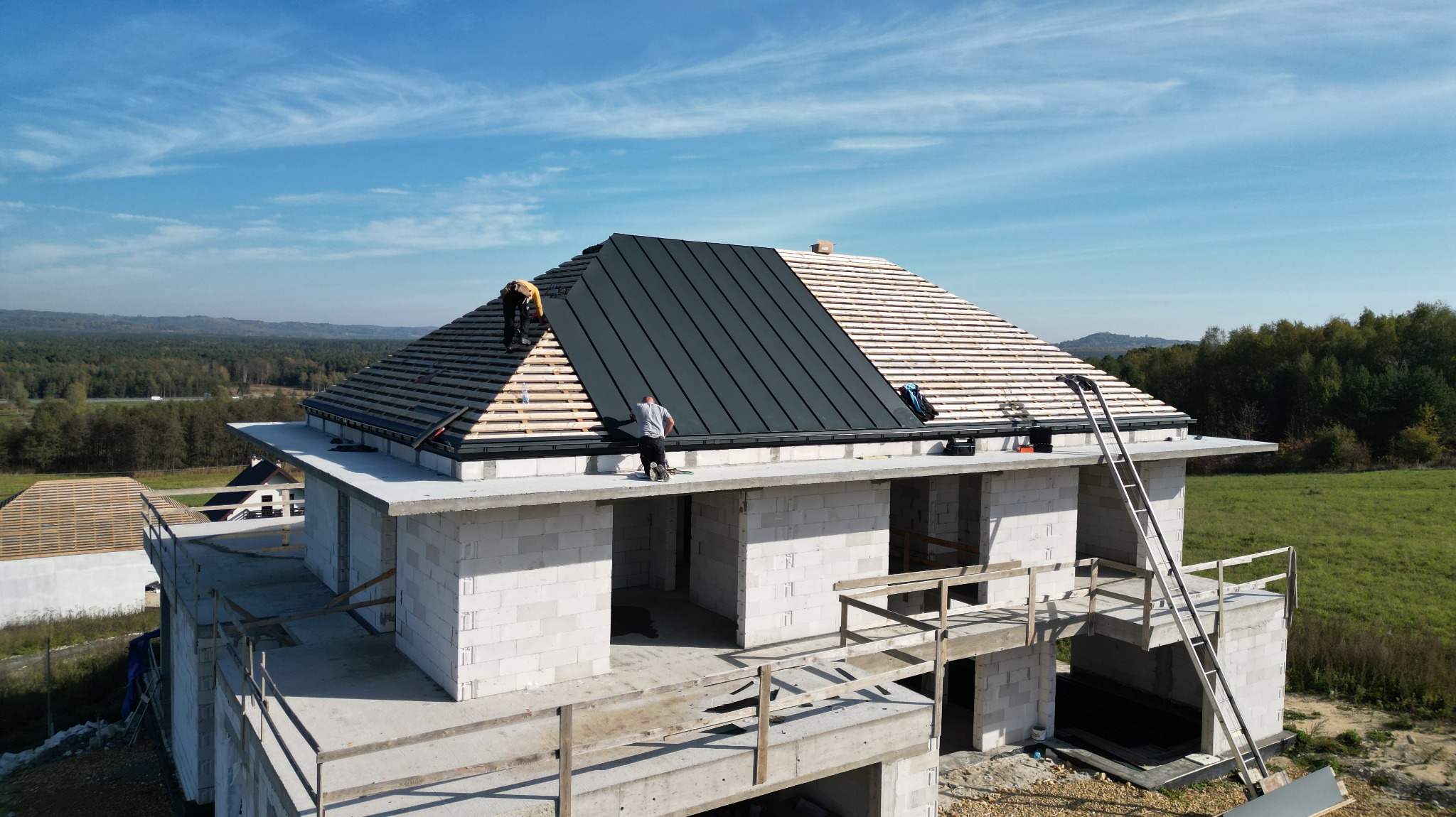 Budowa domu jednorodzinnego: etap montażu czarnego pokrycia dachowego na drewnianej konstrukcji więźby, widoczni dwaj pracownicy na dachu oraz białe ściany z betonu komórkowego parteru, drabina...