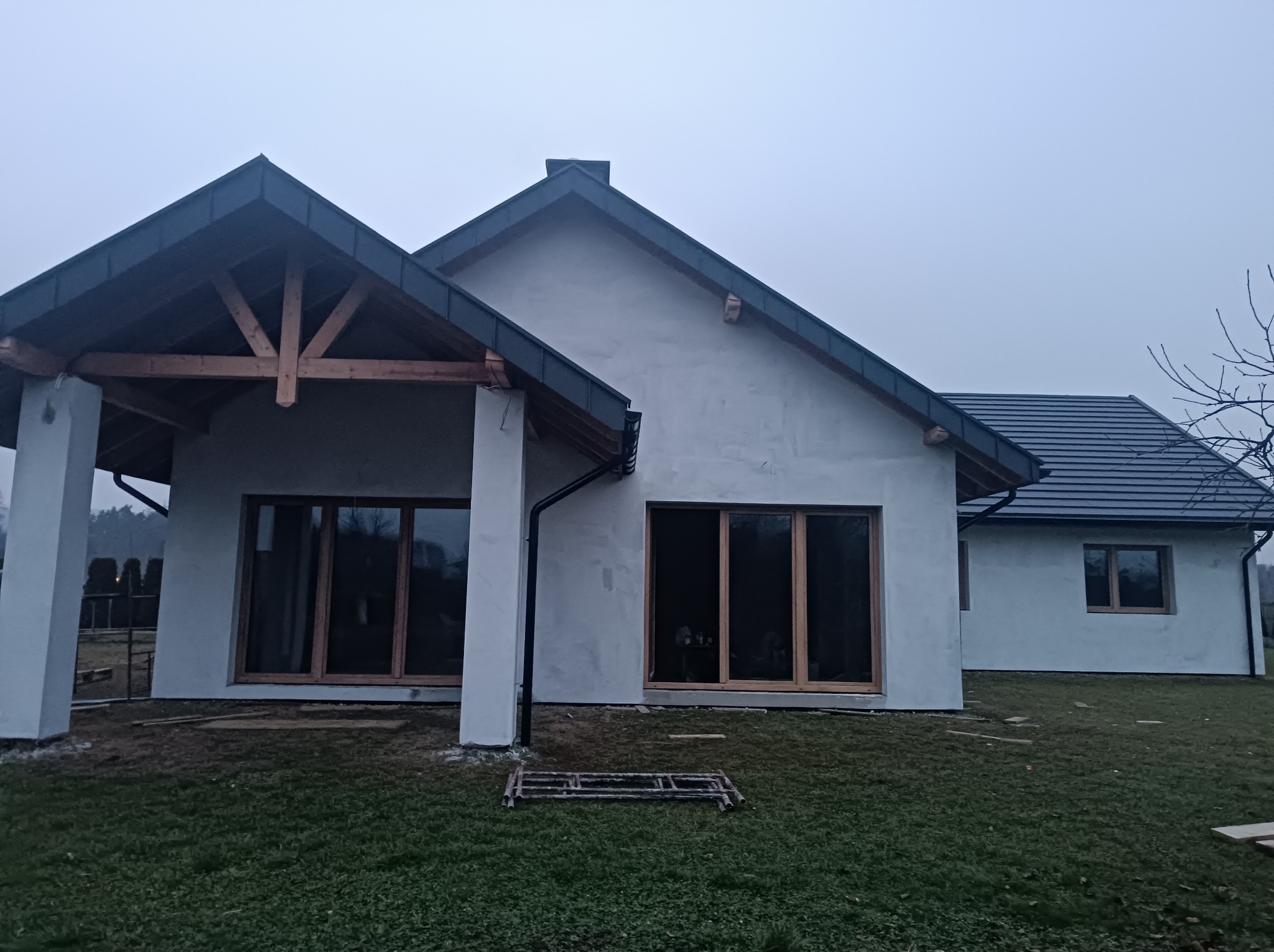 Surowy stan domu jednorodzinnego z widoczną konstrukcją dachu i stolarką okienną, na trawniku leżące rusztowanie, pochmurne niebo w tle.