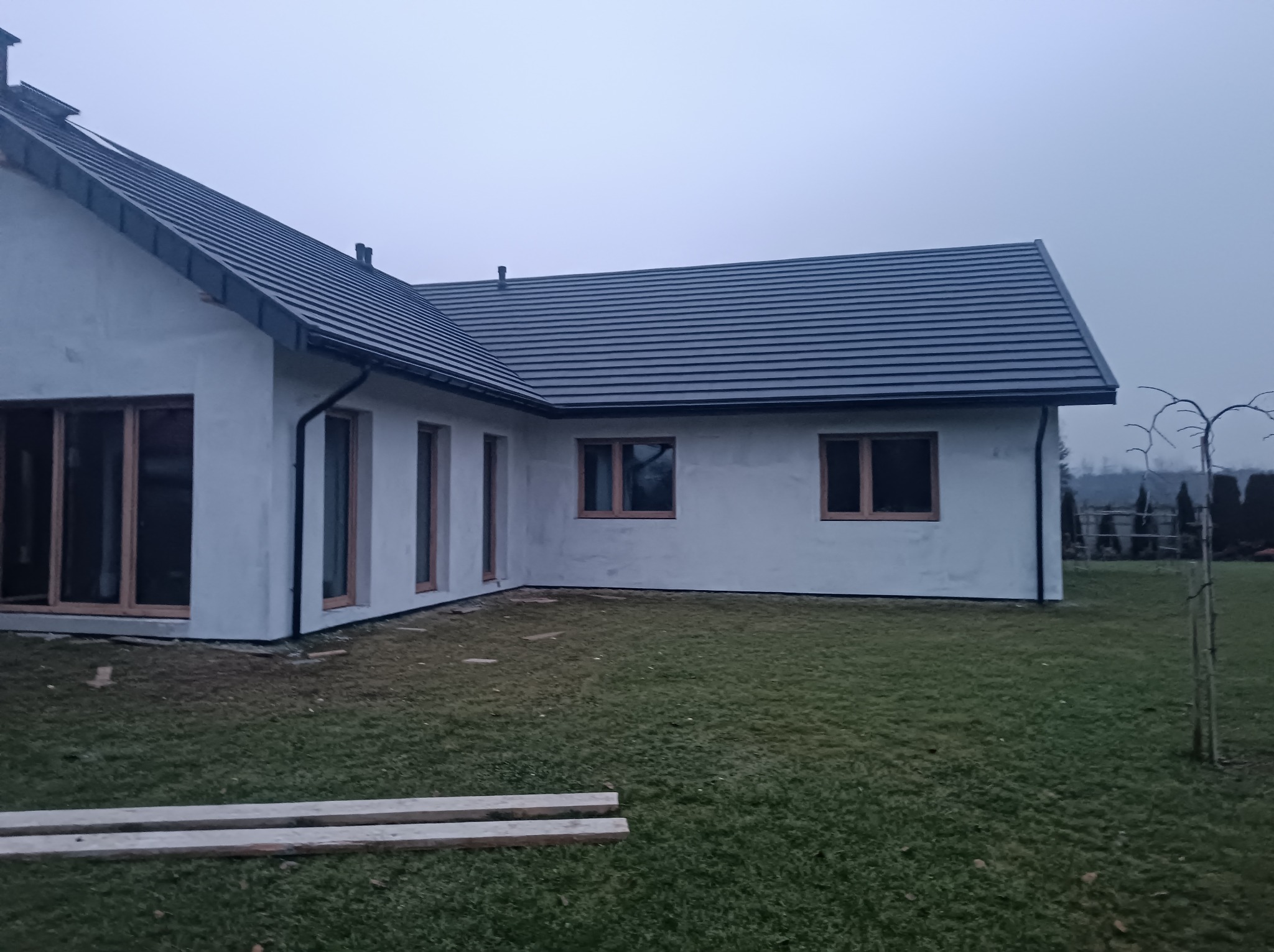 Nowoczesny, parterowy dom z szarą dachówką i drewnianymi ramami okiennymi w trakcie wykończenia elewacji, widok z ogrodu w pochmurny dzień.