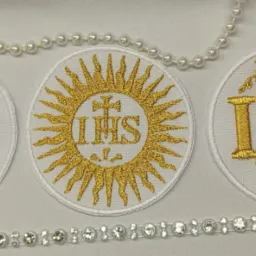 Trzy okrągłe naszywki z haftowanym złotą nicią monogramem IHS, otoczone dekoracyjnymi motywami, leżące na białym tle, z dodatkiem sznurów pereł i kryształków.