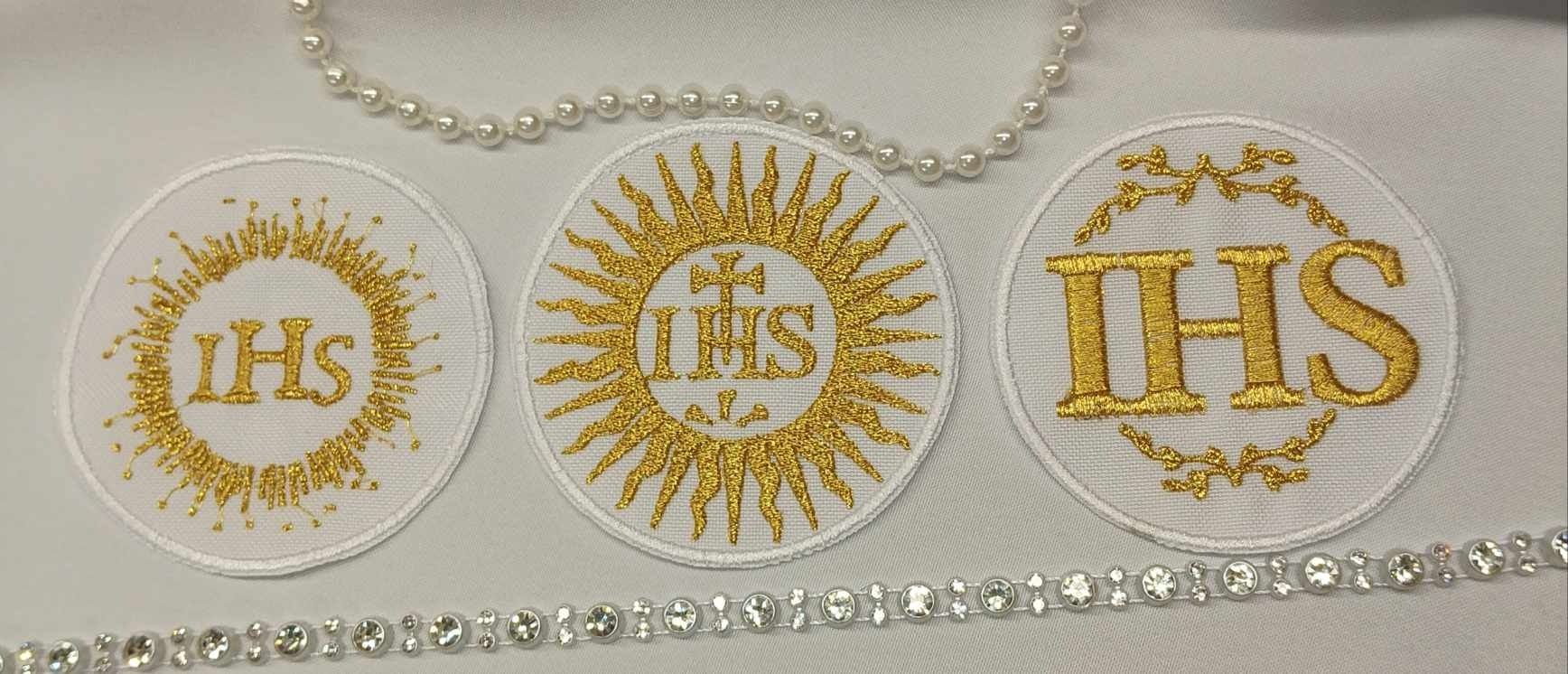 Trzy okrągłe naszywki z haftowanym złotą nicią monogramem IHS, otoczone dekoracyjnymi motywami, leżące na białym tle, z dodatkiem sznurów pereł i kryształków.
